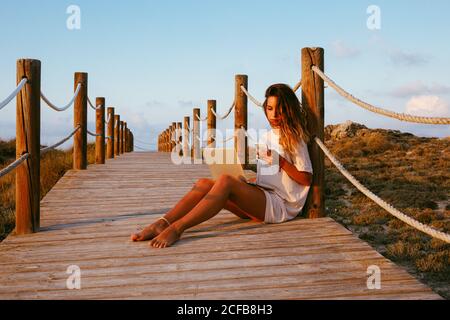 Concentrated female freelancer on vacation in white shirt sitting on empty bridge and working with laptop and using mobile phone on blue sky background Stock Photo