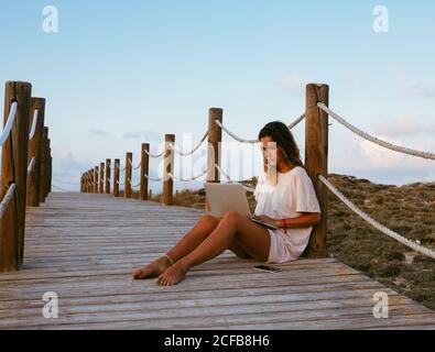 Concentrated female freelancer on vacation in white shirt sitting on empty bridge and working with laptop on blue sky background Stock Photo