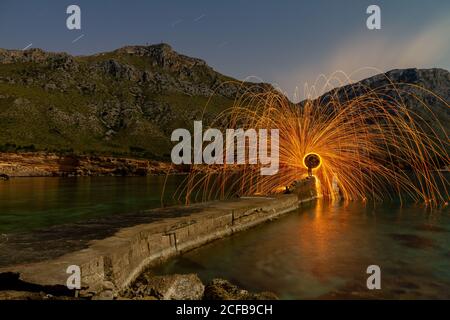 Long exposure of silhouette of human with burning round and fireworks on rocks between water near mountains in evening Stock Photo
