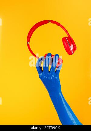 Crop creative person with blue painted hand holding modern headphones on yellow background in studio Stock Photo