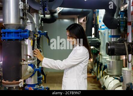 Side view of positive young lady in lab coat checking valves on piping system in industrial facility Stock Photo