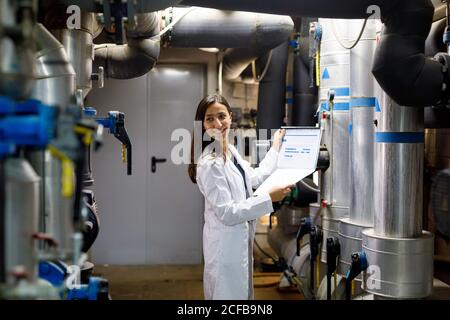 Side view of young Woman in lab coat smiling and looking away while checking data in journal during safety inspection of piping system in industrial facility Stock Photo