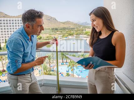 Adult man measuring balcony railing height and young Woman making notes in journal during safety inspection in luxury hotel on resort Stock Photo