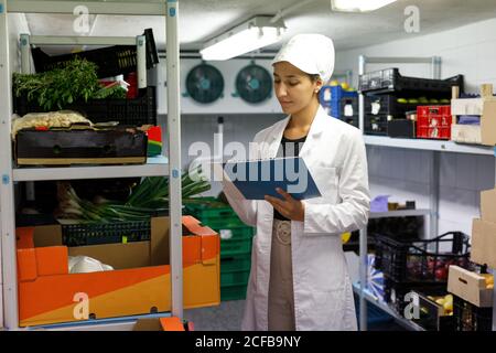 Woman in lab coat and hat making notes while checking food inside cold storage room in cafe Stock Photo