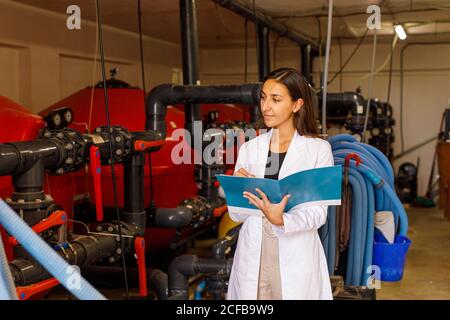 Young female in lab coat checking piping system and writing in journal during safety inspection in industrial facility Stock Photo