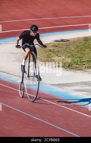 From above adult focused cyclist in black sportswear and helmet riding penny farthing along racetrack at sports stadium while getting ready for competition in sunny day Stock Photo