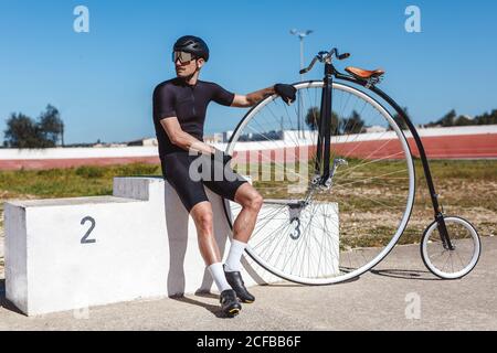 Low angle of serious thoughtful man in black sportswear and helmet looking away and thinking about future competition while sitting alone on champion podium and leaning on retro high wheel bicycle against blurred sports stadium Stock Photo