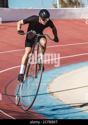 From above adult focused cyclist in black sportswear and helmet riding penny farthing along racetrack at sports stadium while getting ready for competition in sunny day Stock Photo