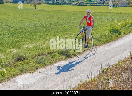 From above adult focused male in sunglasses and stylish casual clothes riding retro high wheel bike along footpath against green grassy field in summer day Stock Photo