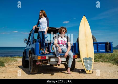 Young energetic man with surfboard and attractive Woman standing on blue offroadster in bright day Stock Photo