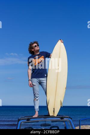 Attractive man with surfboard looking along on roof of blue car in bright day Stock Photo