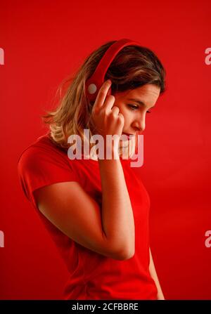 Side view of happy young blonde Woman in vivid headphones and t shirt smiling and looking down while listening to music against red background Stock Photo