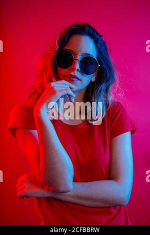 Thoughtful young blonde Woman in round sunglasses touching chin looking at camera while standing against red background Stock Photo