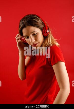 Side view of happy young blonde Woman in vivid headphones and t shirt smiling and looking down while listening to music against red background Stock Photo