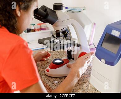 Back view of unrecognizable female doctor looking through modern microscope while at sample while working in modern clinic Stock Photo