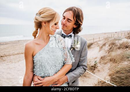 Pleased young groom in wedding suit hugging blonde haired bride in stylish dress behind at empty sandy seashore Stock Photo