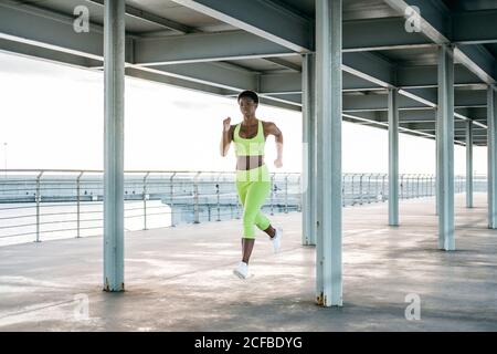 From below African American adult sportsWoman in vibrant green activewear focusing and running alone along waterfront among metal columns under roof Stock Photo