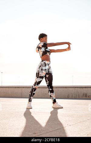 From below focused African American adult Woman in flowered sportswear stretching hand muscles while standing alone and warming up before training among urban environment in sunny day Stock Photo