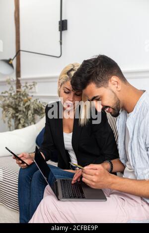 Delighted blonde female smiling and reading credit card credentials to cheerful ethnic boyfriend with laptop while sitting on sofa and making online purchases together Stock Photo