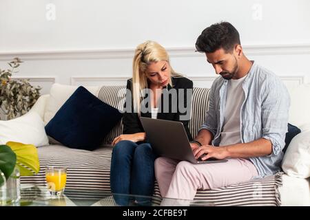 Blonde Woman and ethnic bearded handsome man in casual clothes sitting on sofa and browsing laptop together at home Stock Photo
