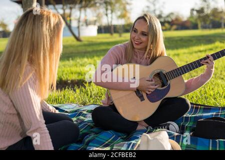 Two blonde girls smiling happily, singing and playing guitar on the grass. Musical picnic in the park during a sunny day. Stock Photo