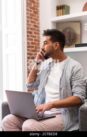 Bearded ethnic man using laptop and speaking on smartphone while working on remote project at home Stock Photo