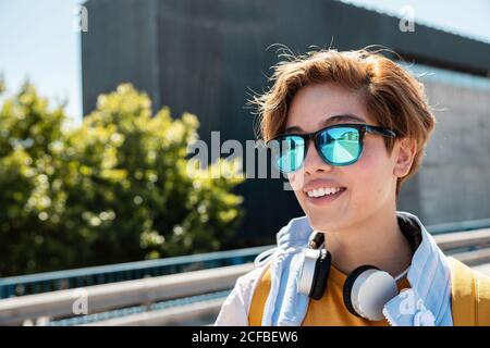 Bright millennial female in stylish sunglasses and vivid white and yellow outfit wearing wireless headphones and enjoying view while looking away against blurred city street Stock Photo
