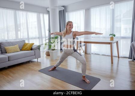 Beautiful active young woman making yoga asana Virabhadrasana warrior 2 pose at home. Healthy lifestyle and sports concept. Series of exercise poses. Stock Photo