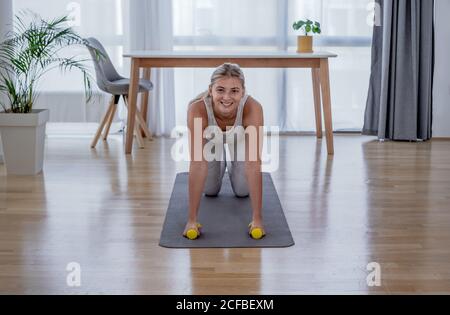 Beautiful active young woman exercise half plank pose with dumbbells at home. Healthy lifestyle and sports concept. Series of exercise poses. Stock Photo