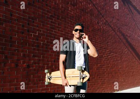 From below young African American cheerful handsome man in casual clothes and stylish sunglasses with long board speaking on phone Stock Photo