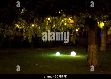 illumination backyard light garden with electric ground lantern with round diffuser lamp and garland of light bulbs on tree branches with leaves, land Stock Photo
