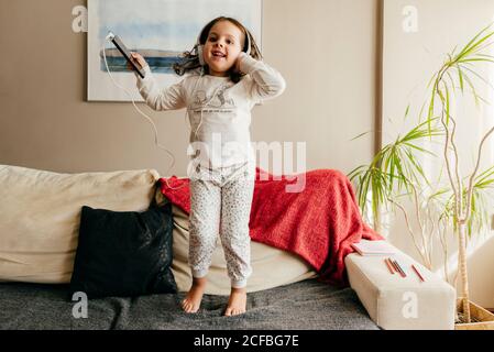 Full length excited little girl in sleepwear and headphones listening to music on smartphone and jumping on soft sofa while having fun at home Stock Photo