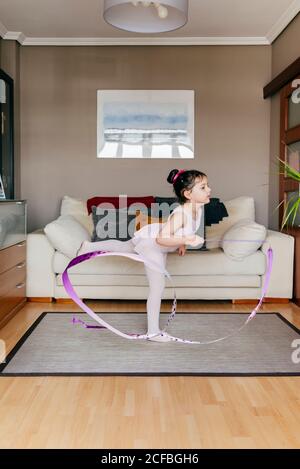 Cute little girl in leotard and tights spinning ribbon and dancing during rhythmic gymnastic training in cozy living room at home Stock Photo