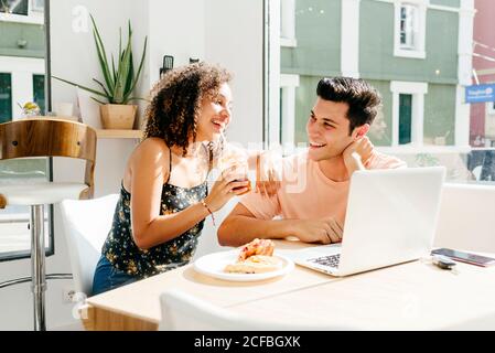 Cheerful ethnic Woman enjoying fresh beverage and leaning on shoulder of boyfriend with laptop while sitting at cafe table Stock Photo