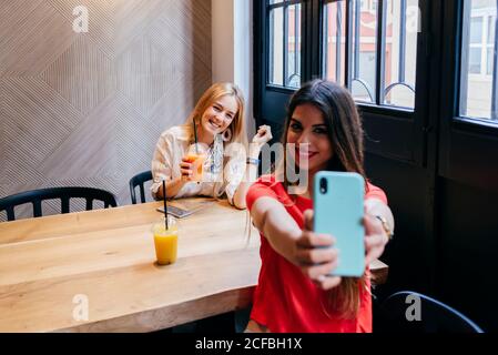 Pretty happy young female friends having meeting in cafe and taking selfie with smartphone while sitting at wooden table and drinking fruit juice Stock Photo