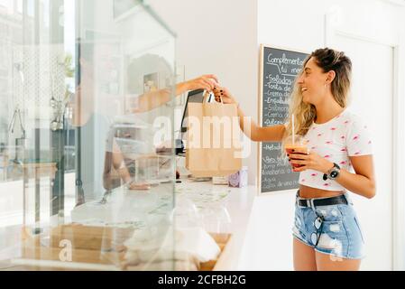 Happy young female with healthy drink taking paper bag with order from bartender while standing near counter in restaurant Stock Photo