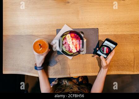 Top view of crop Woman holding disposable cup of orange juice and checking picture of food on smartphone while sitting at wooden table with fruit bowl Stock Photo