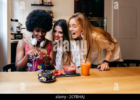 Happy young diverse female friends watching funny video on smartphone while spending morning in small cafe and having healthy breakfast together Stock Photo