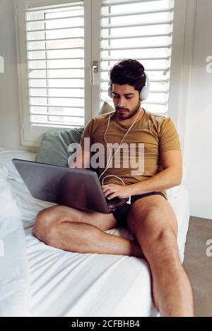 Young concentrated successful man in casual shirt and headphones sitting on bed using a laptop computer Stock Photo