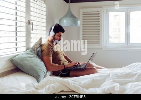 Young concentrated successful man in casual shirt and headphones sitting on bed using a laptop computer Stock Photo