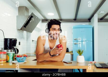 Young smiling handsome male with stylish hairstyle in headset leaning on table in kitchen and holding ripe apple Stock Photo