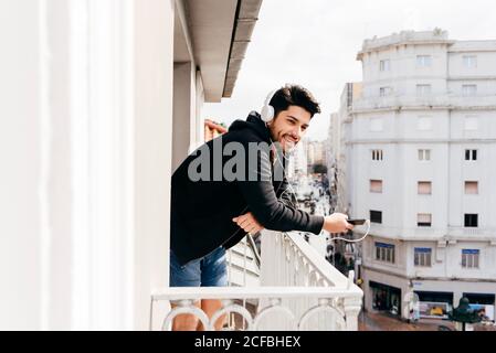Young smiling handsome casual male in headset leaning on terrace railing with smartphone and looking away Stock Photo