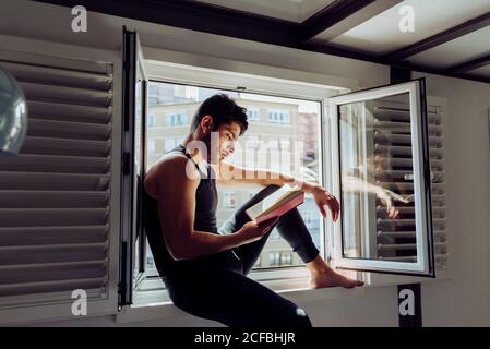 Side view of young serious handsome man in casual sleeveless shirt sitting on window and reading interesting book Stock Photo