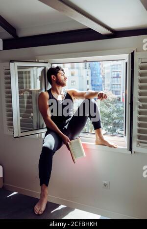 Side view of young serious handsome man in casual sleeveless shirt sitting on window and reading interesting book Stock Photo