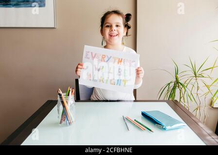 girl sitting at wooden table with colored pencils and demonstrating drawing saying Everything will be alright at camera Stock Photo