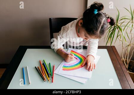 From above of anonymous girl in casual clothes sitting on chair at wooden table and drawing colorful rainbow with pencil on paper Stock Photo