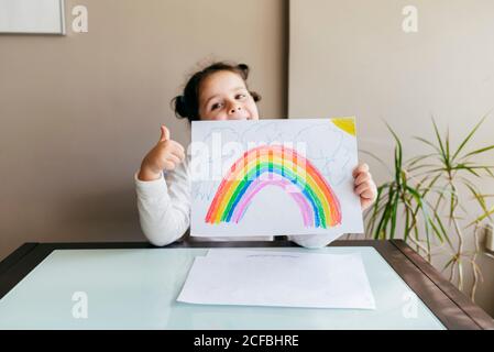 Smiling charming girl holding sheet of paper with colorful drawing while sitting at wooden table and smiling and looking at camera Stock Photo
