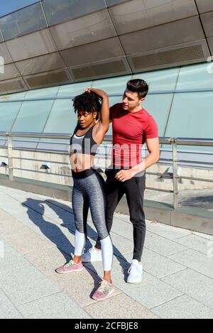 Young athletic man in red shirt giving support to active African American female in performing of stretching exercise during workout in sunny day in city Stock Photo