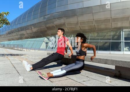 Side view of African American sportsWoman in black sportswear and sportsman in red shirt doing push up exercises next to concrete bench in city Stock Photo