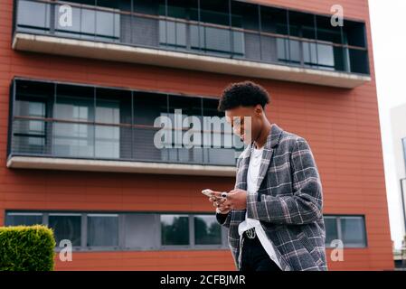 From below side view of happy young African American male student in stylish outfit browsing smartphone while standing on city street near modern building Stock Photo